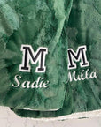 A green double-sided plush blanket with satin cording finish, featuring custom text 'Sadie' and 'Milla' along with the letter 'M' in bold font.