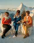 Three women sitting in the snow with a mountainous background, enjoying drinks.

