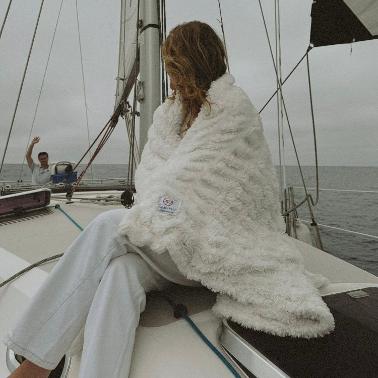 A person sitting on a boat deck wearing a white fluffy blanket with embroidery, with the ocean and another person in the background.