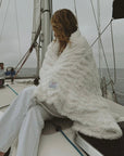 A person sitting on a boat deck wearing a white fluffy blanket with embroidery, with the ocean and another person in the background.