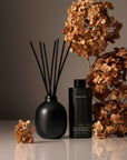 Black diffuser with reeds and a bottle of Home&Garden candle in front of dried flowers on a reflective surface.